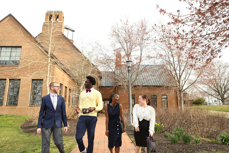 Four people dressed in business attire walk and talk outside a brick building on a cloudy day, surrounded by early spring trees and landscaping.