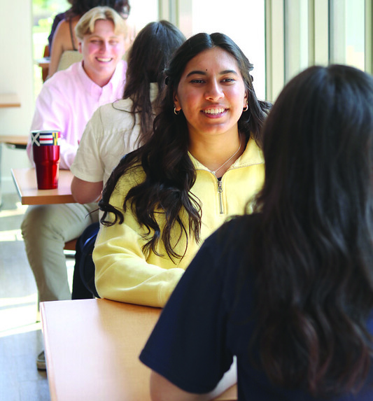 Several students sit in pairs at tables by a large window, engaged in conversation inside a well-lit room.