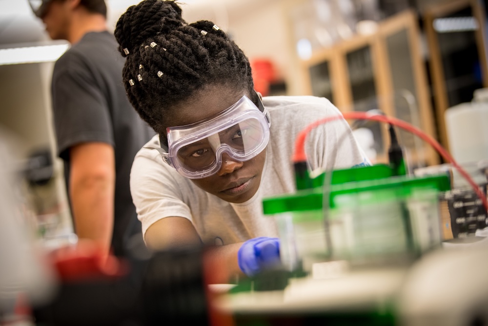 A person wearing safety goggles and gloves closely observes an experiment in a laboratory setting with scientific equipment on the table.