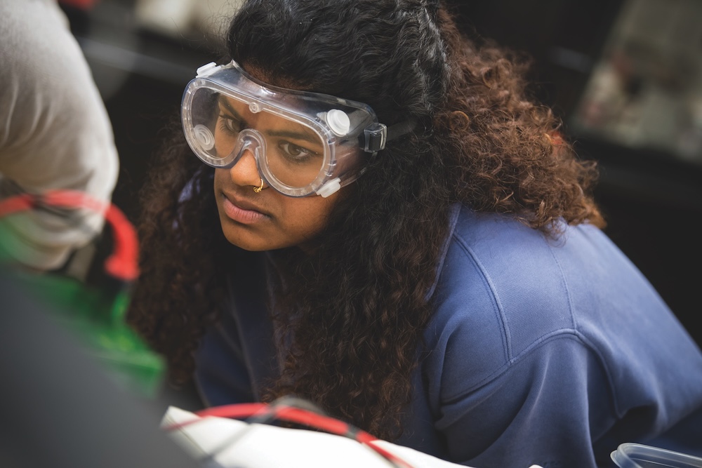 Person wearing safety goggles and a blue sweatshirt closely observes something in a laboratory or classroom setting.