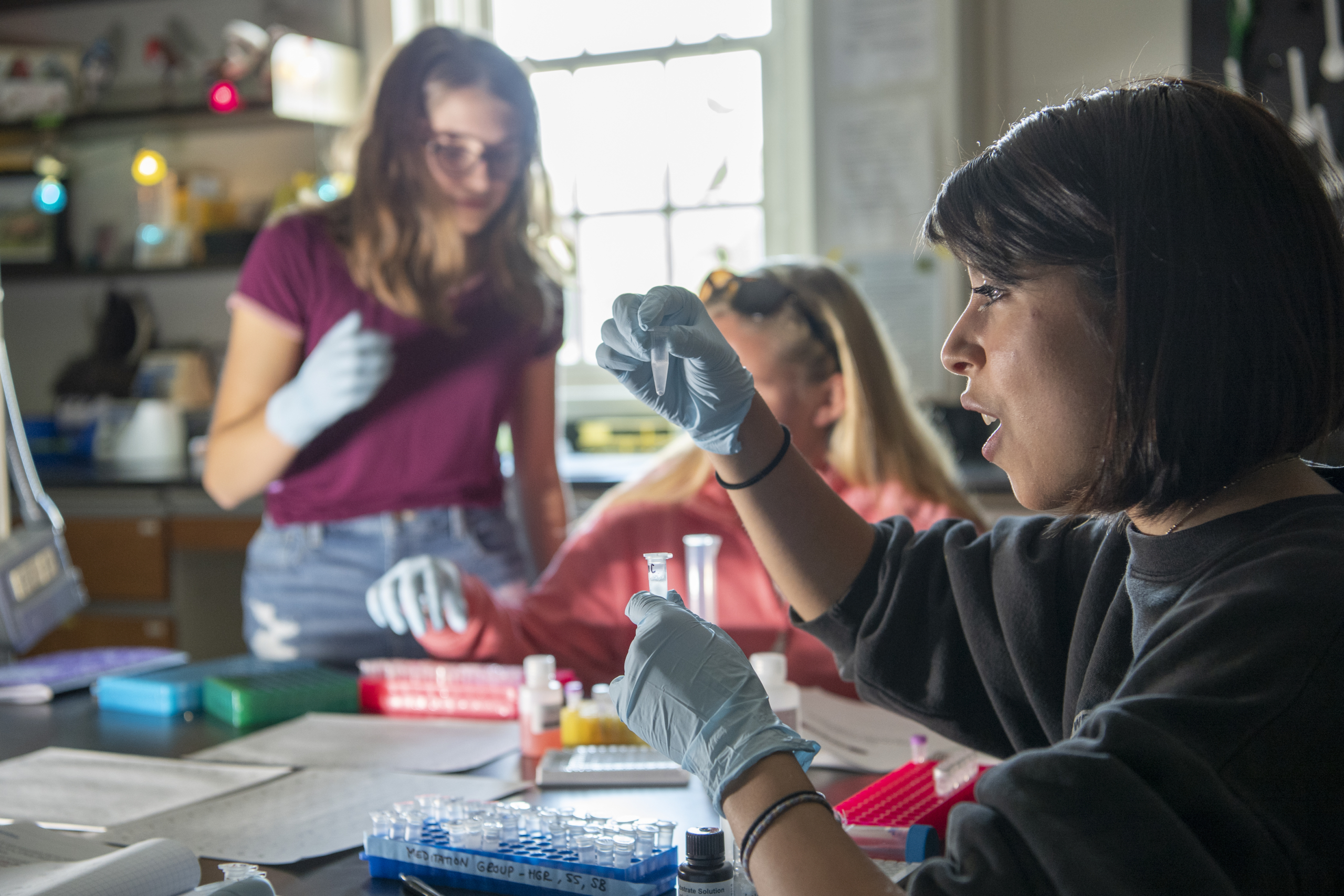 Three people wearing gloves work with test tubes and lab equipment at a table in a brightly lit laboratory.