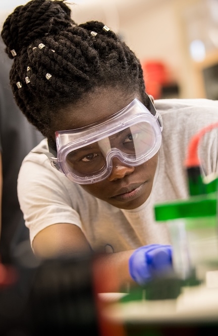 A person wearing safety goggles and gloves closely observes a laboratory experiment involving scientific equipment.