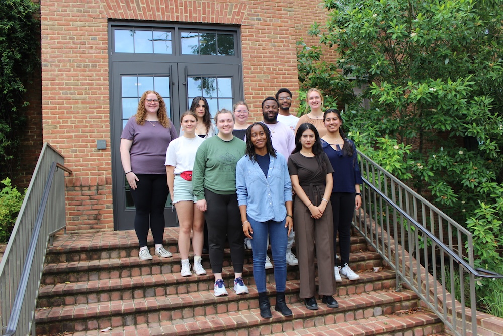 A group of ten people pose together on outdoor brick steps in front of a building with glass doors and greenery on the side.