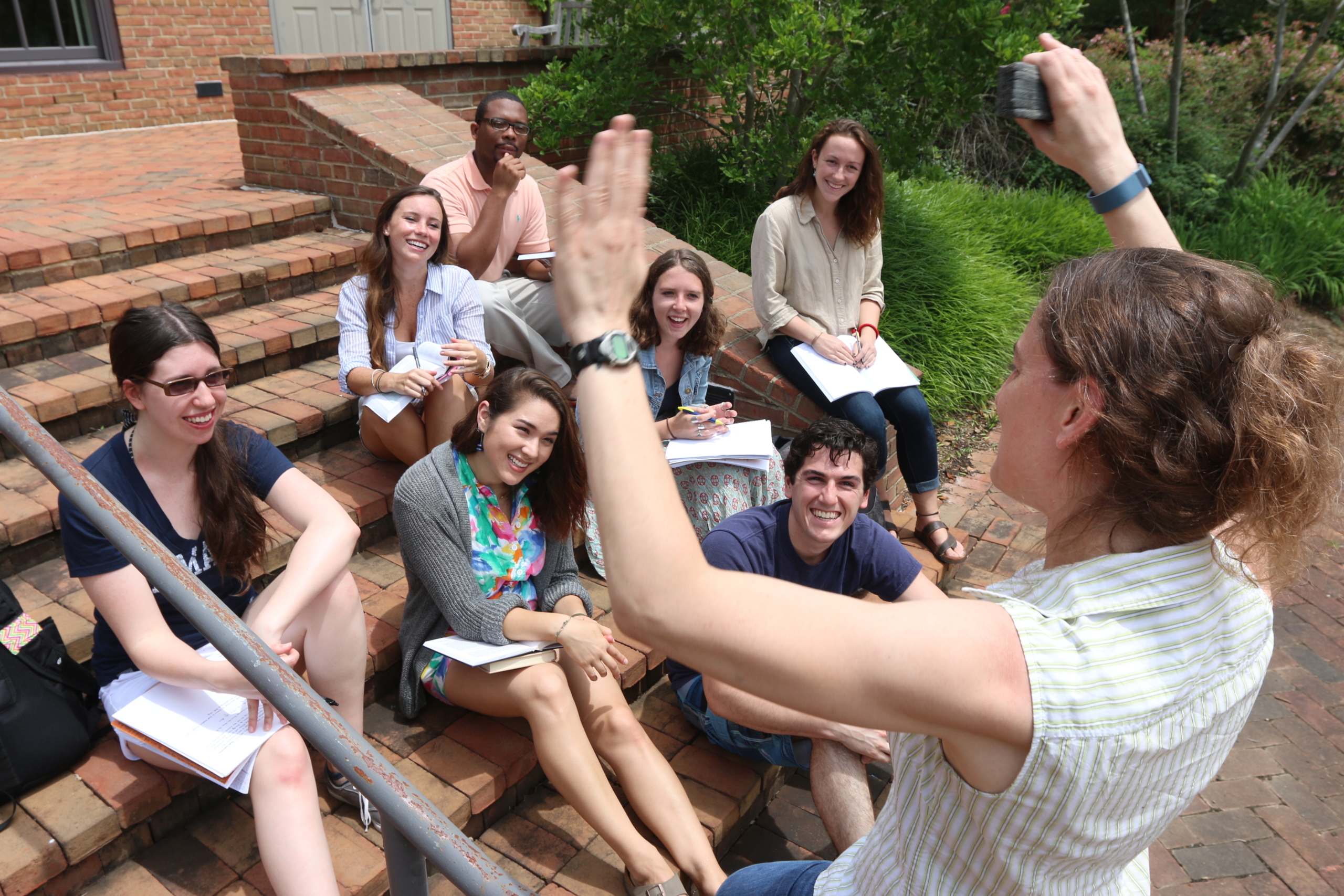 A group of students sit on outdoor brick steps, smiling and watching a woman standing in front of them raising her hand as she speaks. Some students hold notebooks.