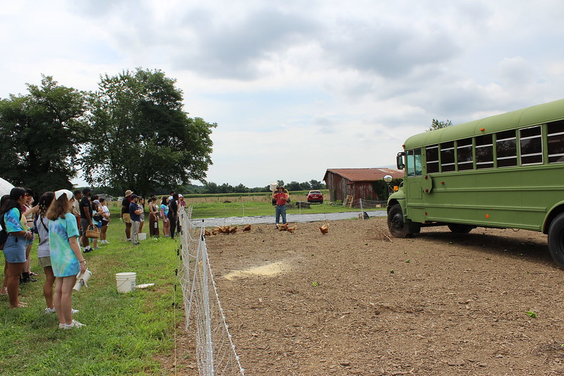 A group of people stand near a fenced area with chickens, watching a person speak next to a green school bus on a farm under a cloudy sky.