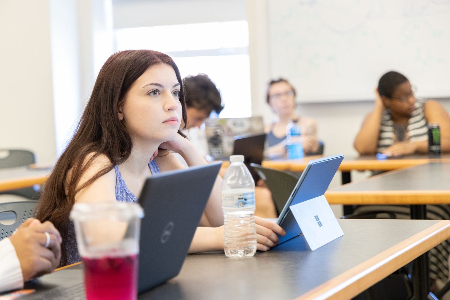 Students sit at desks in a classroom, some using laptops and tablets. A woman in the foreground listens attentively with a water bottle and a tablet in front of her.