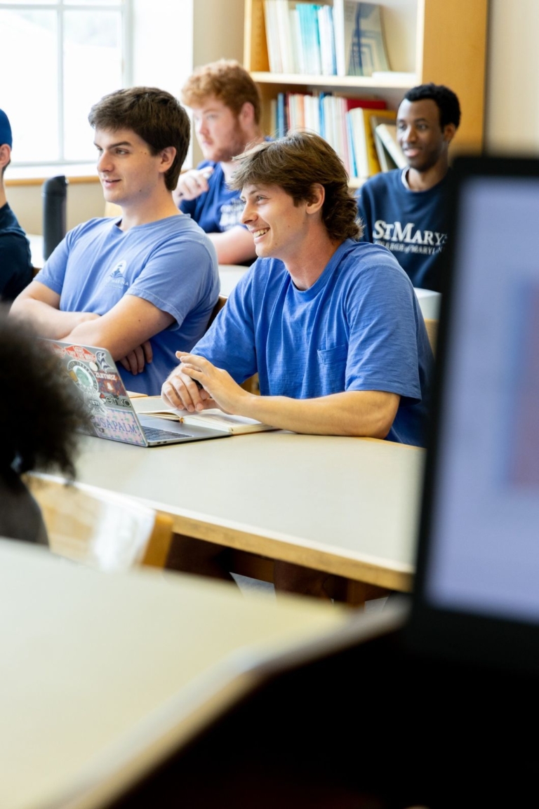 Several students sit at desks in a classroom, some smiling and engaged, with laptops and notebooks open in front of them. Bookshelves and a window are in the background.