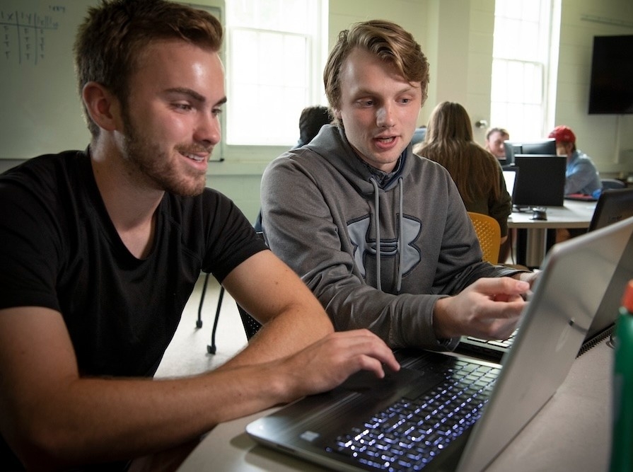 Two young men sit at a table working together on laptops in a classroom setting, with other students and computers visible in the background.