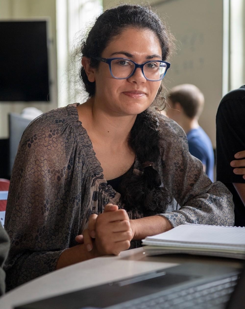 A person with glasses and dark hair sits at a table with a notebook, looking at the camera. Other people and a whiteboard are visible in the background.