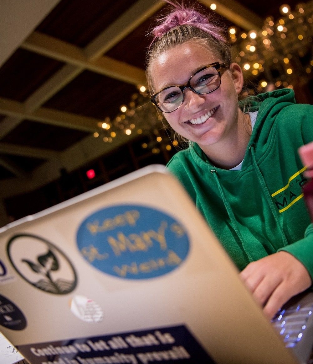 A person wearing glasses and a green hoodie smiles while sitting at a table, working on a laptop decorated with various stickers.