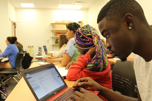 A student works on a laptop in a classroom while others sit at desks, some writing in notebooks, facing the front of the room.