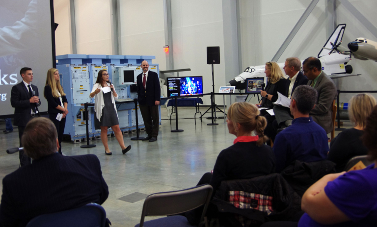 A group of people present and listen in a hangar-like room with aerospace equipment and a drone on display in the background.