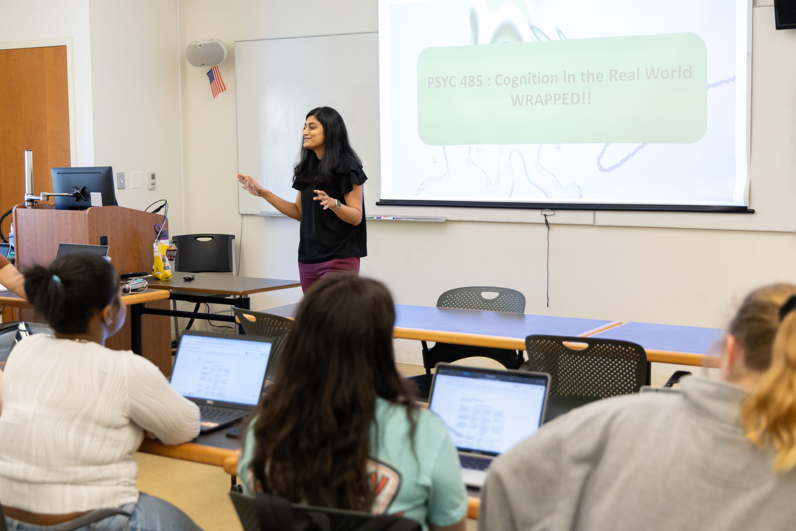 A woman stands and speaks at the front of a classroom, presenting a slide titled "PSYC 485: Cognition in the Real World WRAPPED!" to students seated with laptops.