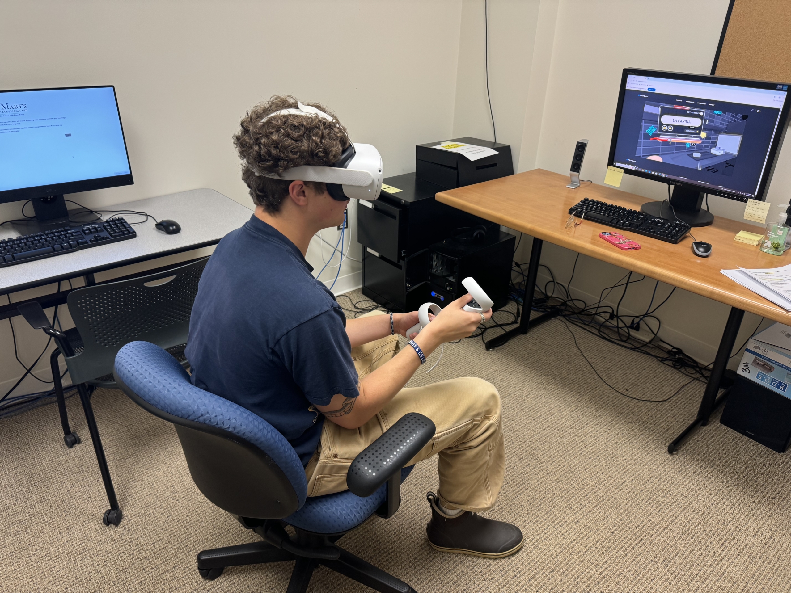 A person wearing a VR headset and holding controllers is seated in an office with multiple desks, computers, and a monitor displaying a 3D interface.