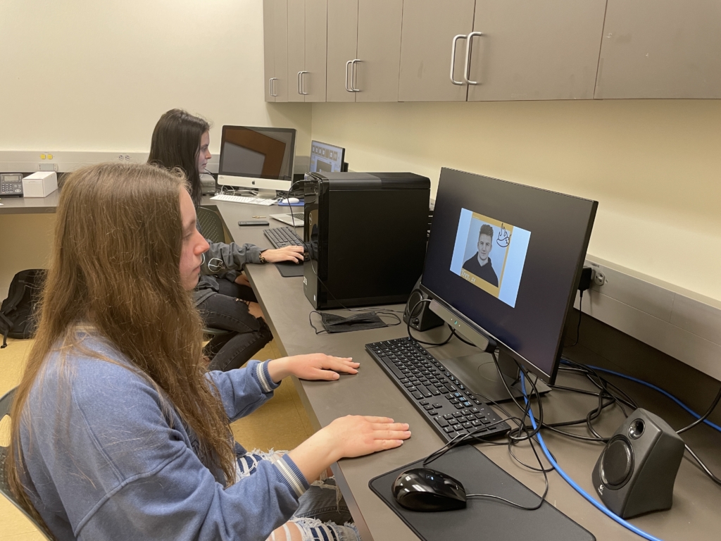Two people sit at desks using desktop computers in a classroom. One monitor displays a person speaking on a video call or online class.