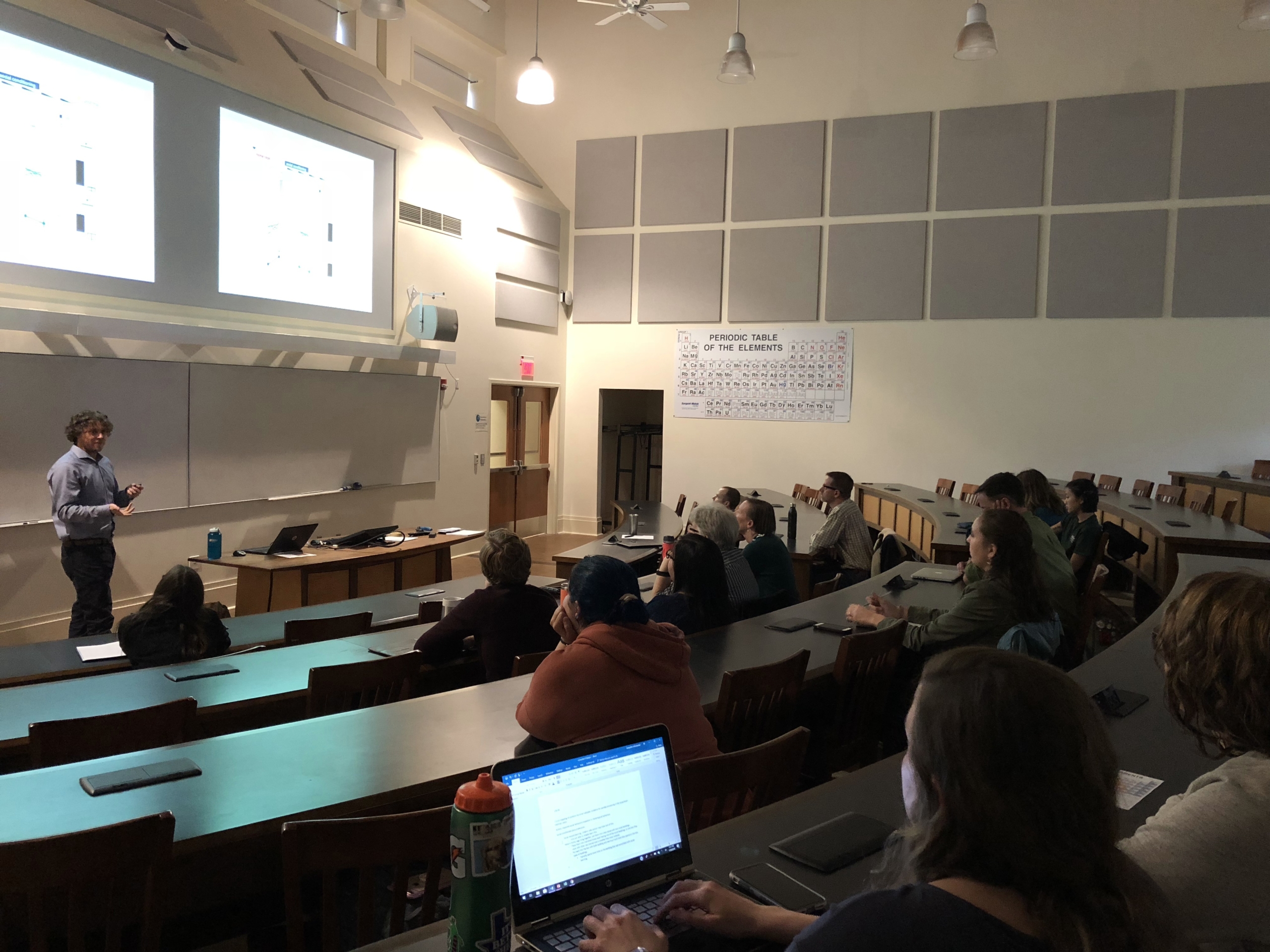 A professor presents slides to students in a college lecture hall. Some students take notes on laptops, while others listen. A periodic table is displayed on the wall.