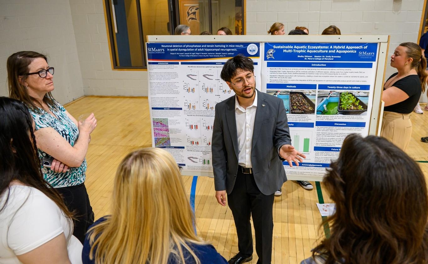 A man in a suit presents research on a poster to a small group of people in a gymnasium setting. Other posters and attendees are visible in the background.