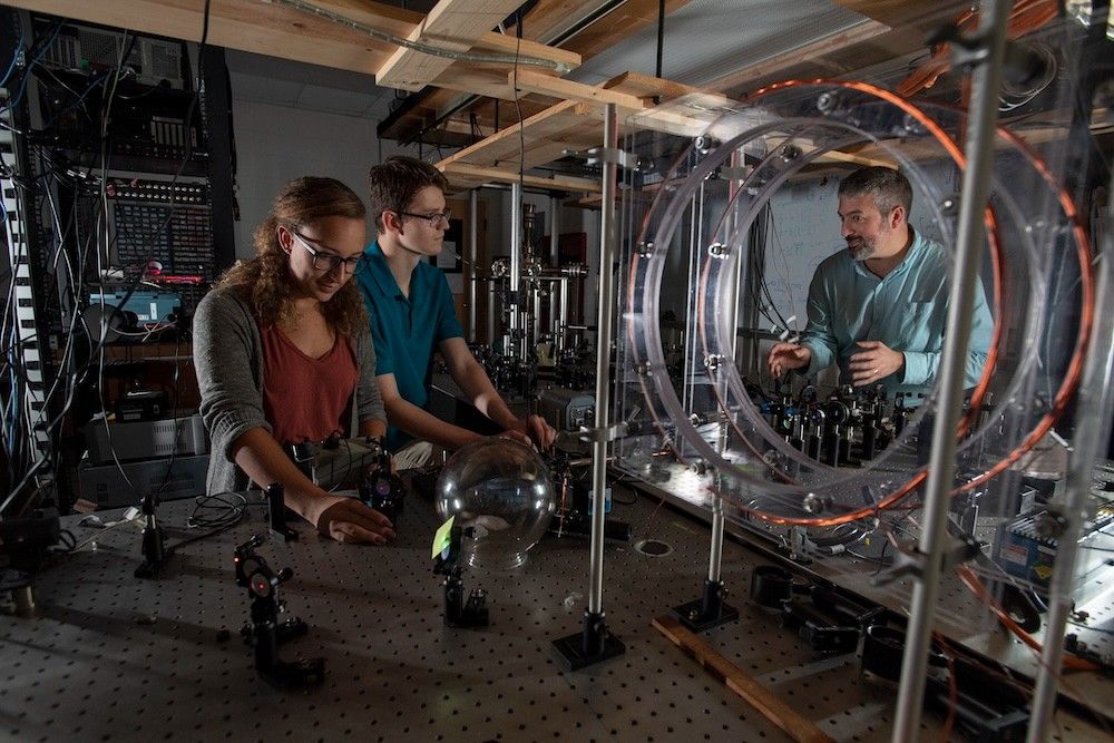 Three people stand around a lab bench with scientific equipment and optical devices, discussing an experiment in a laboratory setting.