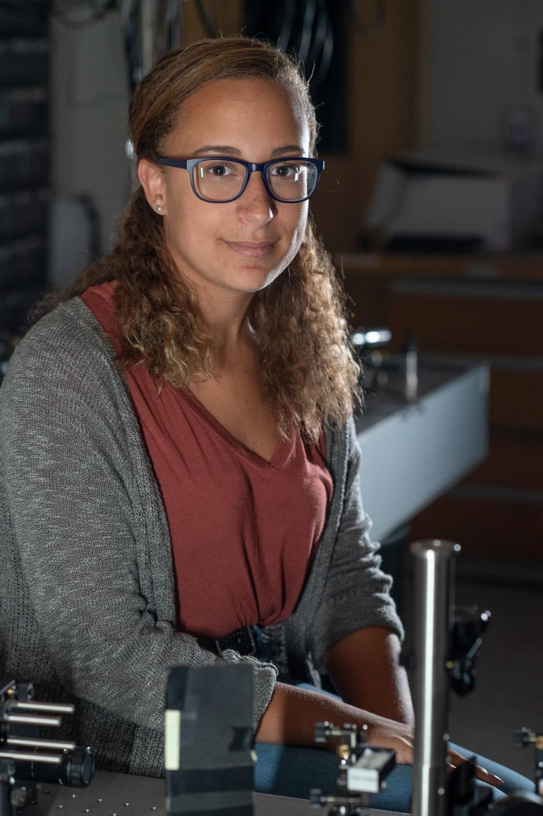 A woman with glasses and curly hair sits at a lab workstation with scientific equipment in a dimly lit room.