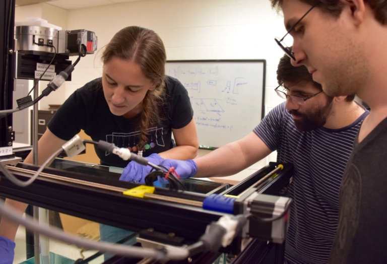 Three people work together on a piece of laboratory equipment, with one adjusting controls while others observe; a whiteboard with diagrams is visible in the background.