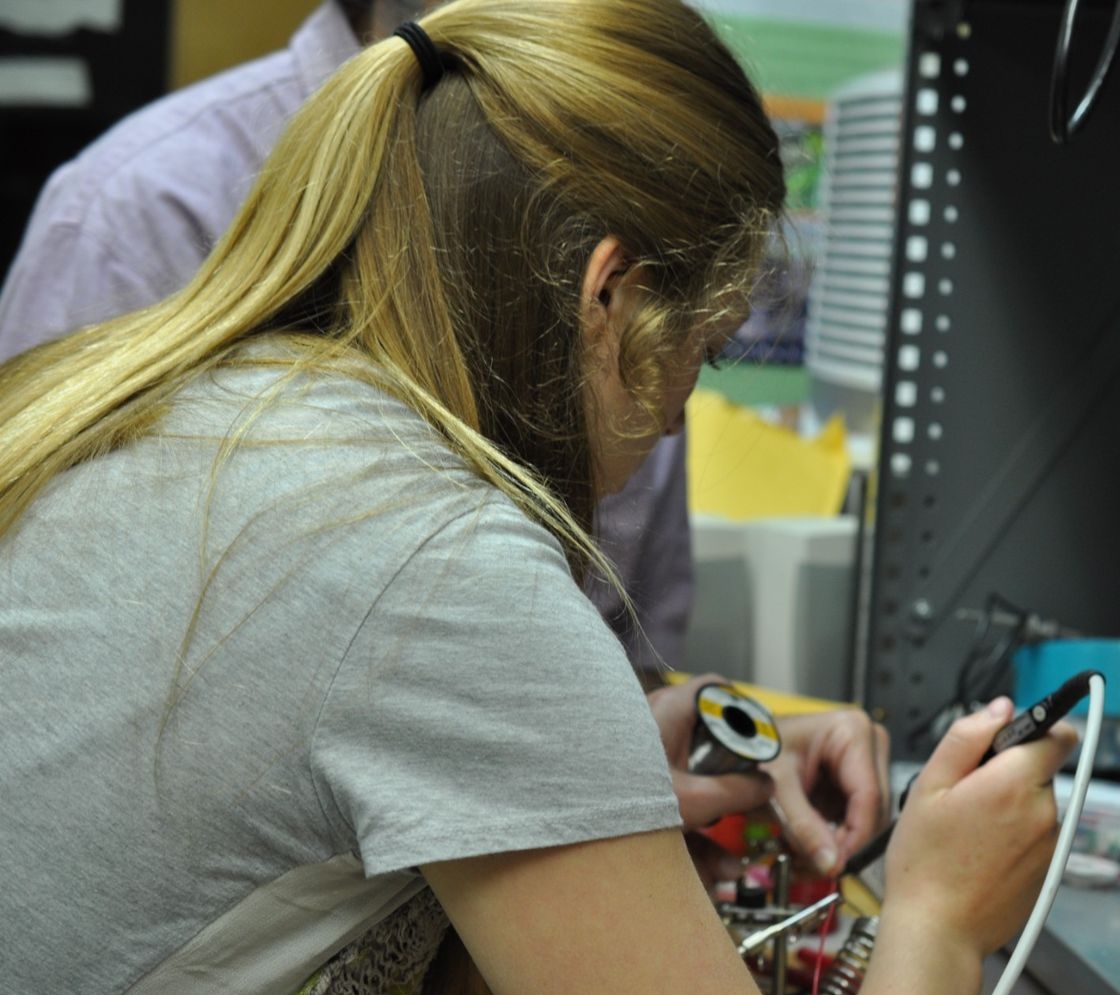 A person with blonde hair soldering electronic components at a workbench, holding a soldering iron and solder wire.