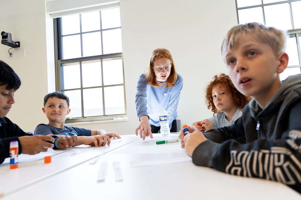 A teacher stands at a table with four students who are seated, interacting with learning materials in a bright classroom.