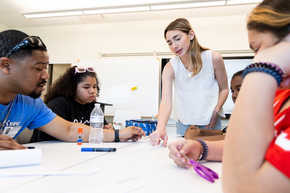 A teacher stands and points at materials on a table while four students sit and work on an activity in a classroom.