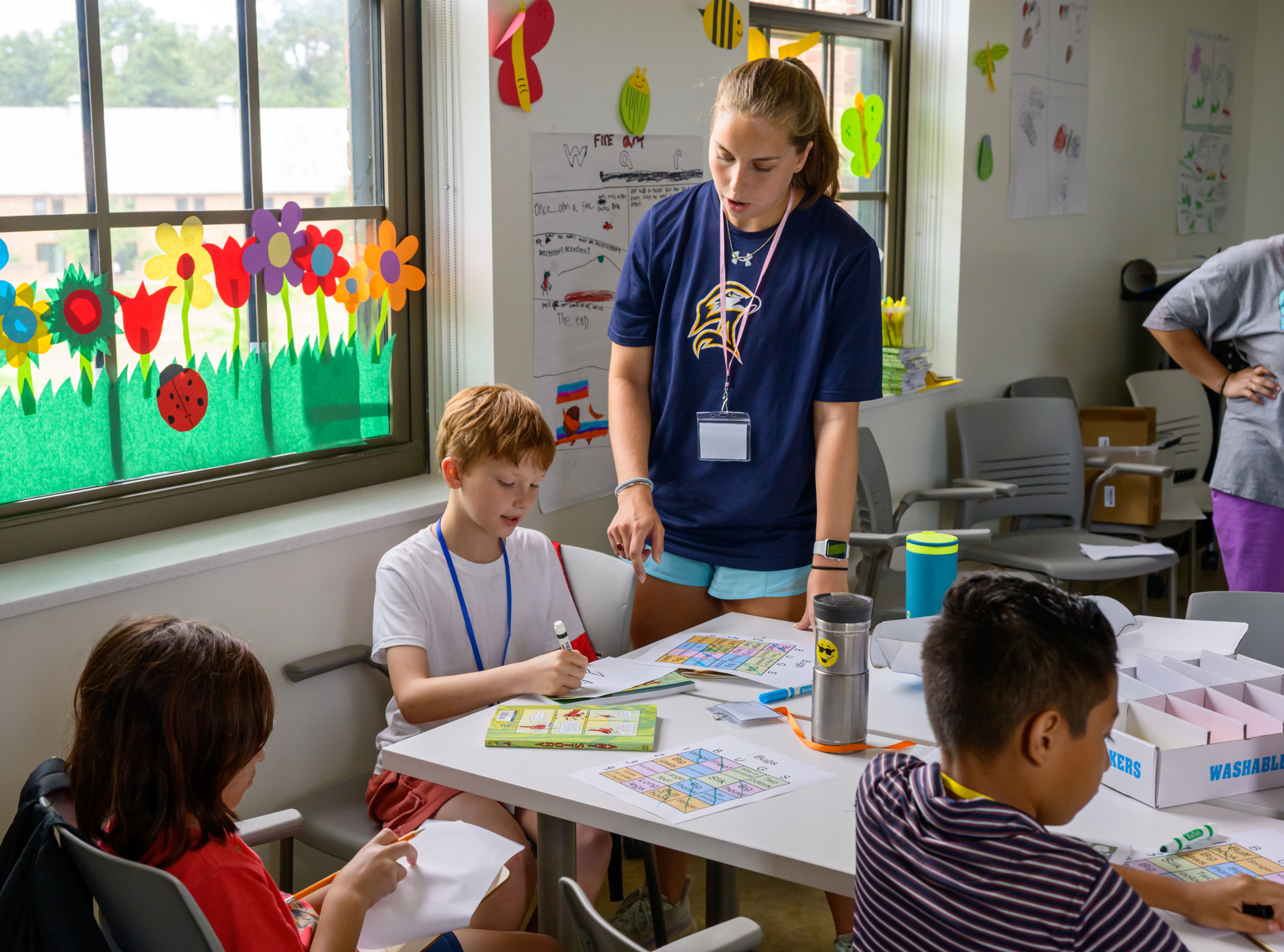 A teacher stands next to three children seated at a table, helping them with art and coloring activities in a classroom decorated with flowers and drawings.