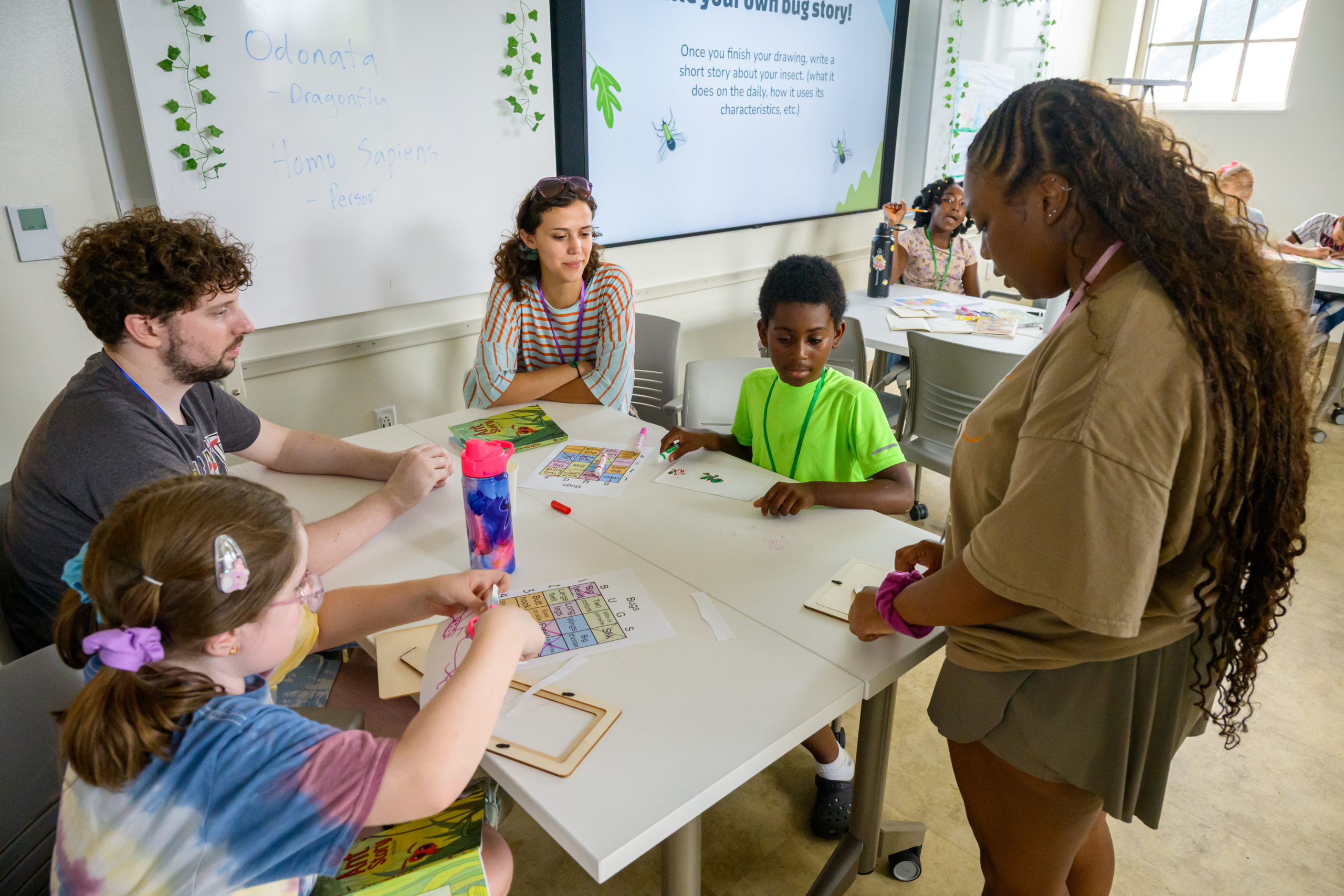A teacher stands with three children at a classroom table working on an activity, while another adult helps a fourth child nearby; a whiteboard with instructions is in the background.
