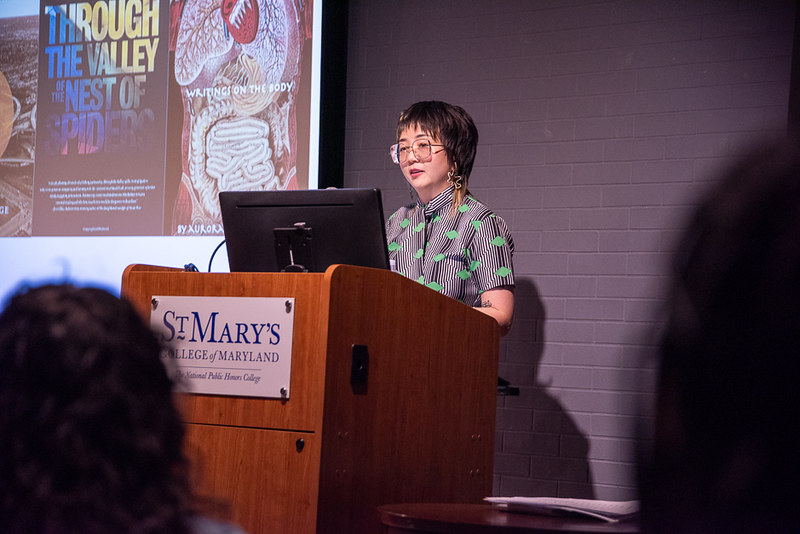 A person stands at a podium labeled "St. Mary's College of Maryland" giving a presentation with book covers displayed on the screen behind them.