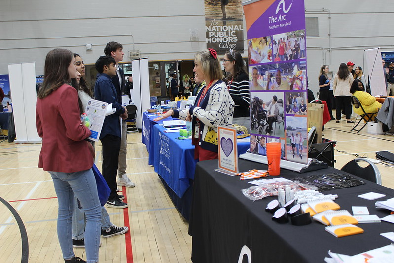 People interact with representatives at informational booths in a gymnasium during an event or fair, with brochures and promotional items on display.