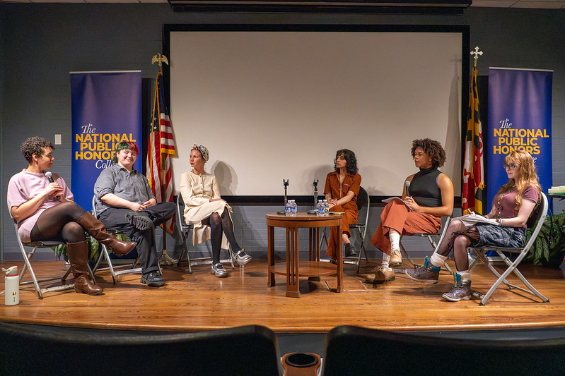 Six people sit in a panel discussion on stage in front of a screen, flanked by two "National Public 荣誉学院" banners and flags.