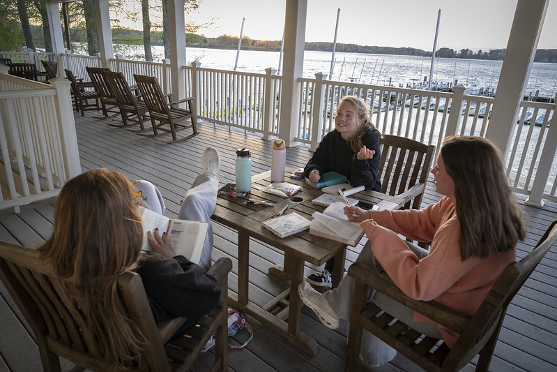 Three young women sit on a porch by the water, reading books and studying at a wooden table with notebooks and water bottles.