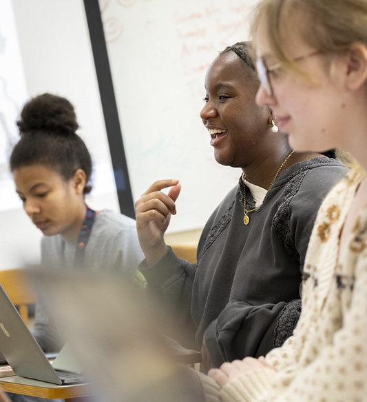 Three students sit at a table working on laptops, engaged in conversation in a classroom setting.