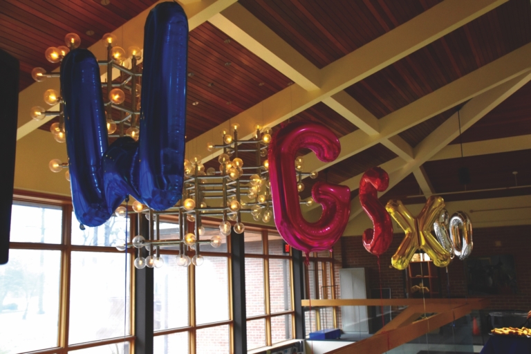Large metallic balloons in blue, red, and gold spelling "WGxO" hang from a ceiling with modern light fixtures in a room with wooden and glass architectural details.