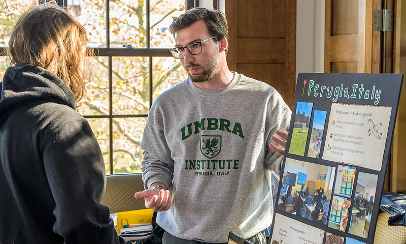 A man in an "UMBRIA INSTITUTE" sweatshirt discusses a display board about Perugia, Italy with another person at an indoor event.