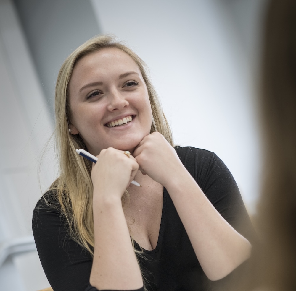 A woman with long blonde hair sits at a table, smiling and holding a pen, resting her chin on her hands while listening to someone off camera.