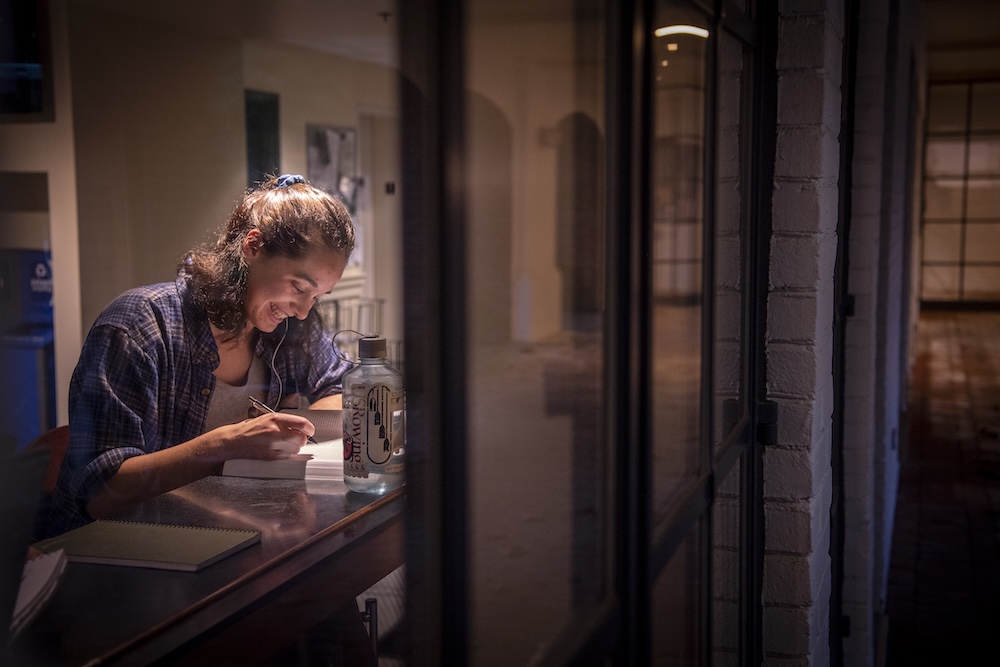 A woman sits alone at a table by a window, writing in a notebook under indoor lighting, with a water bottle and another notebook beside her.