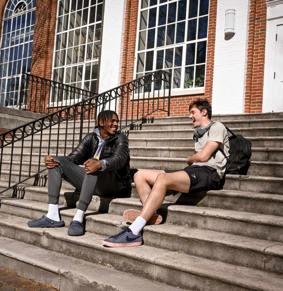Two young men sit on outdoor steps in front of a brick building, talking and laughing. One wears a jacket and pants; the other wears a T-shirt, shorts, and has headphones around his neck.