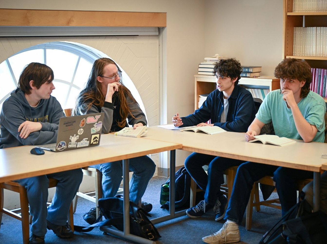 Four students sit around a table in a library or classroom, discussing books open in front of them; one has a laptop with stickers on it.