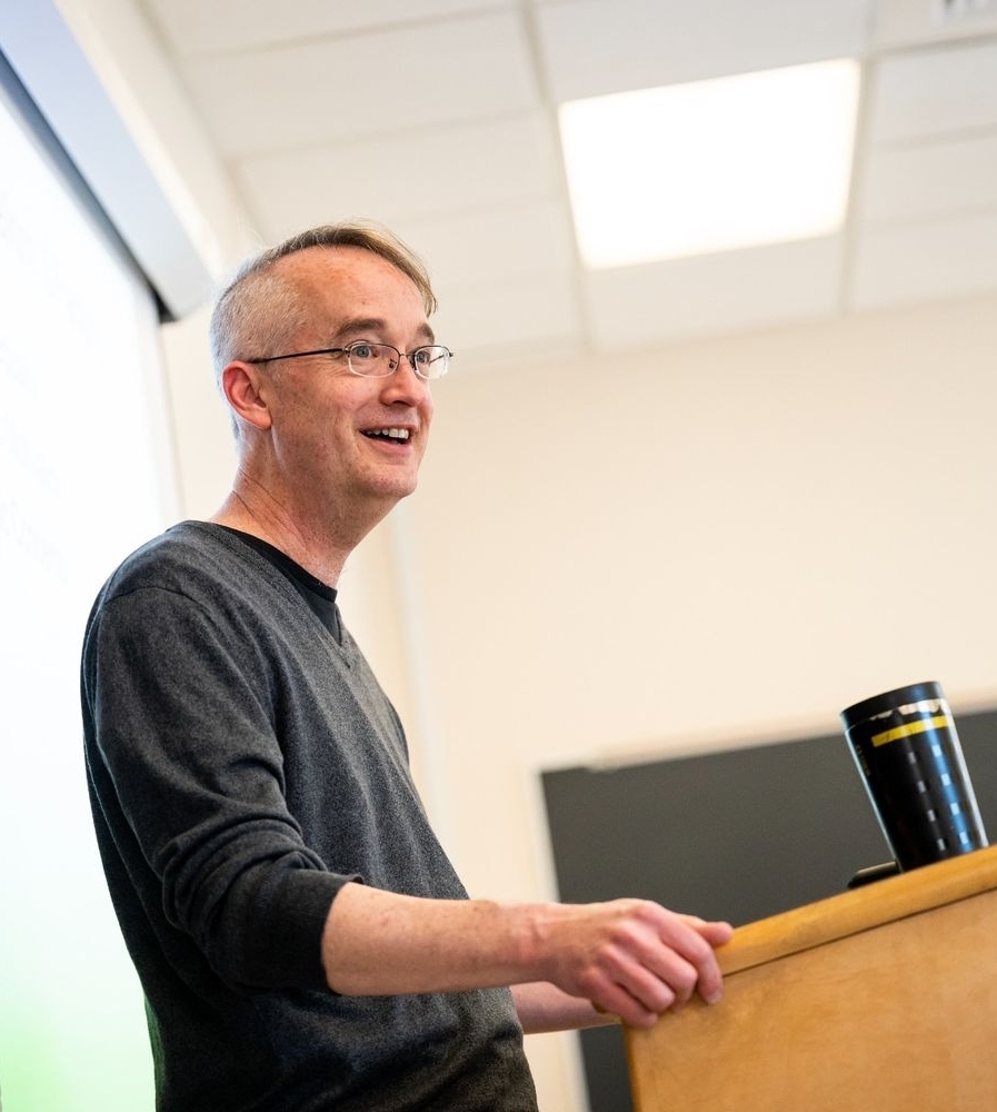 A man wearing glasses stands at a lectern, speaking in a classroom with a projection screen and chalkboard in the background.