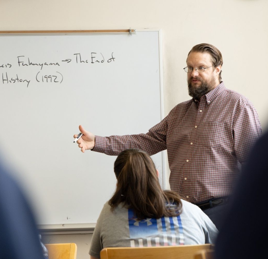 A man stands in front of a whiteboard giving a lecture to students. The whiteboard has the text “Francis Fukuyama → The End of 历史 (1992)” written on it.