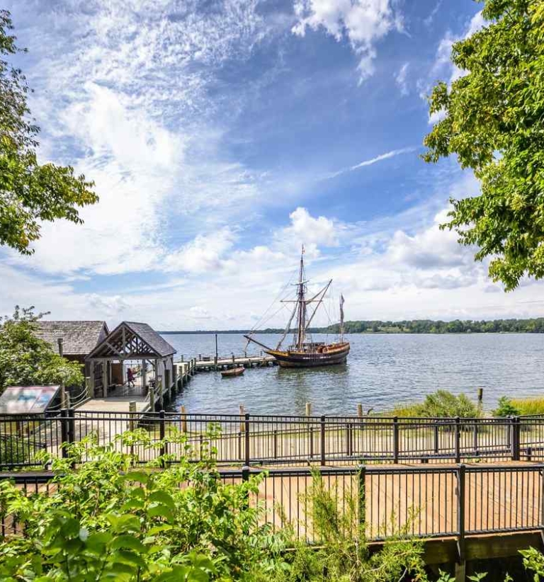 A wooden sailing ship is docked at a pier on a wide river, with trees, a footbridge, and a small building in the foreground under a partly cloudy sky.