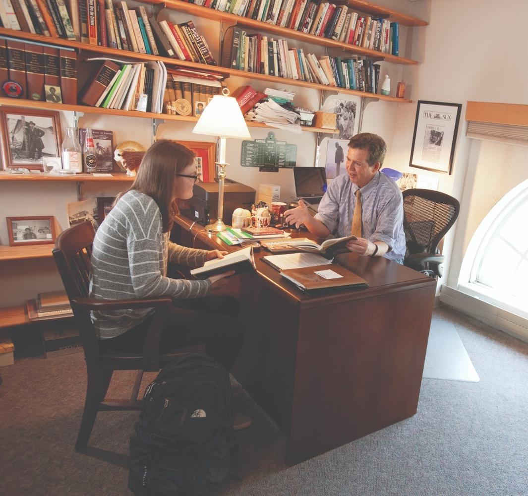 A student sits across from a professor in a book-filled office, discussing papers at his desk near a window.