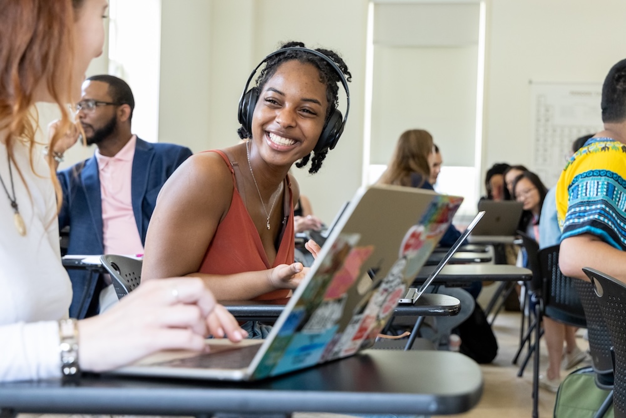 A woman wearing headphones smiles at a classmate while sitting at a desk with a laptop in a classroom setting.