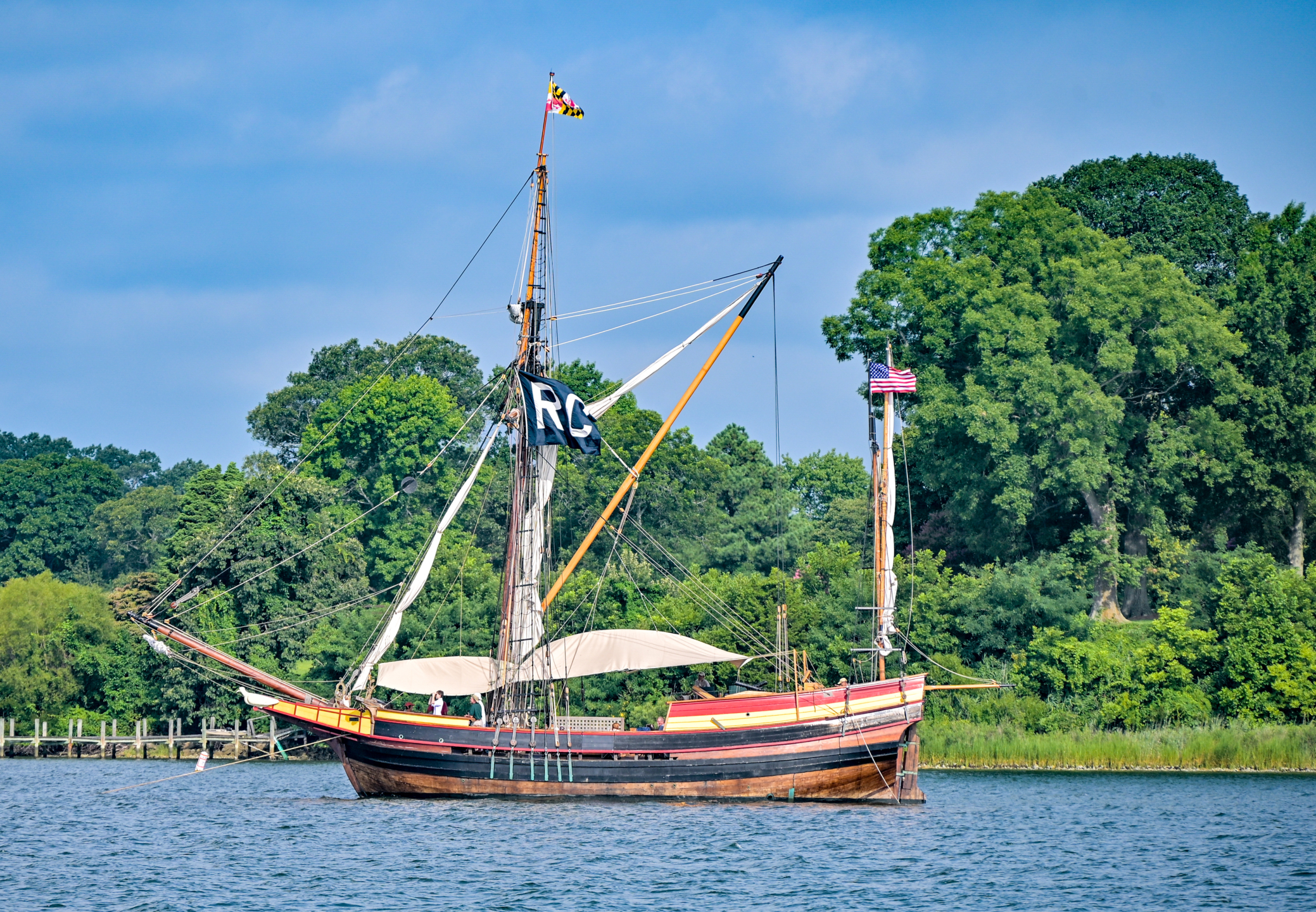 A wooden sailing ship with a black pirate flag is anchored on a calm river, surrounded by green trees and a partly cloudy blue sky.