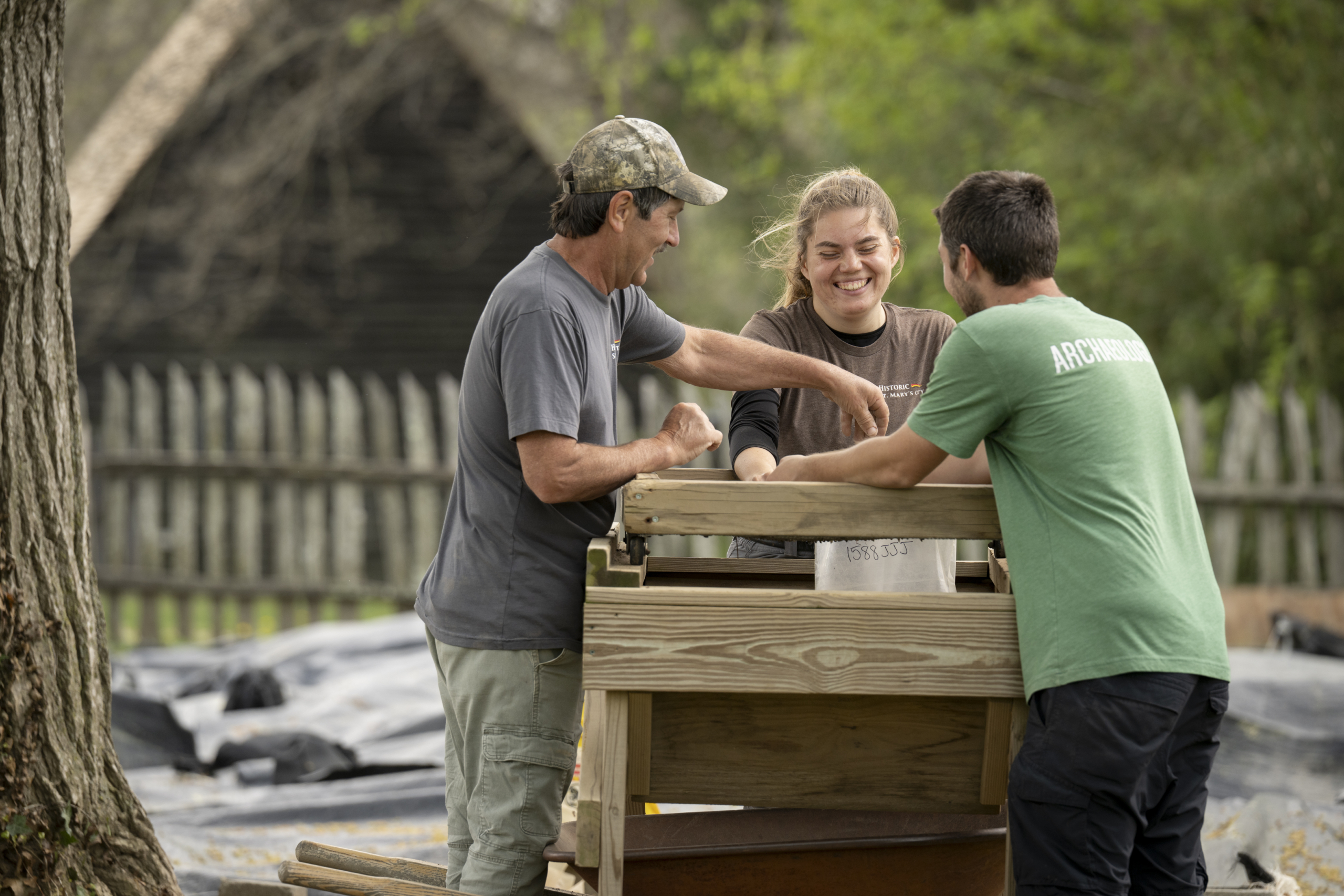 Three people stand outdoors and sift soil using a wooden screen, with trees and a wooden fence in the background.