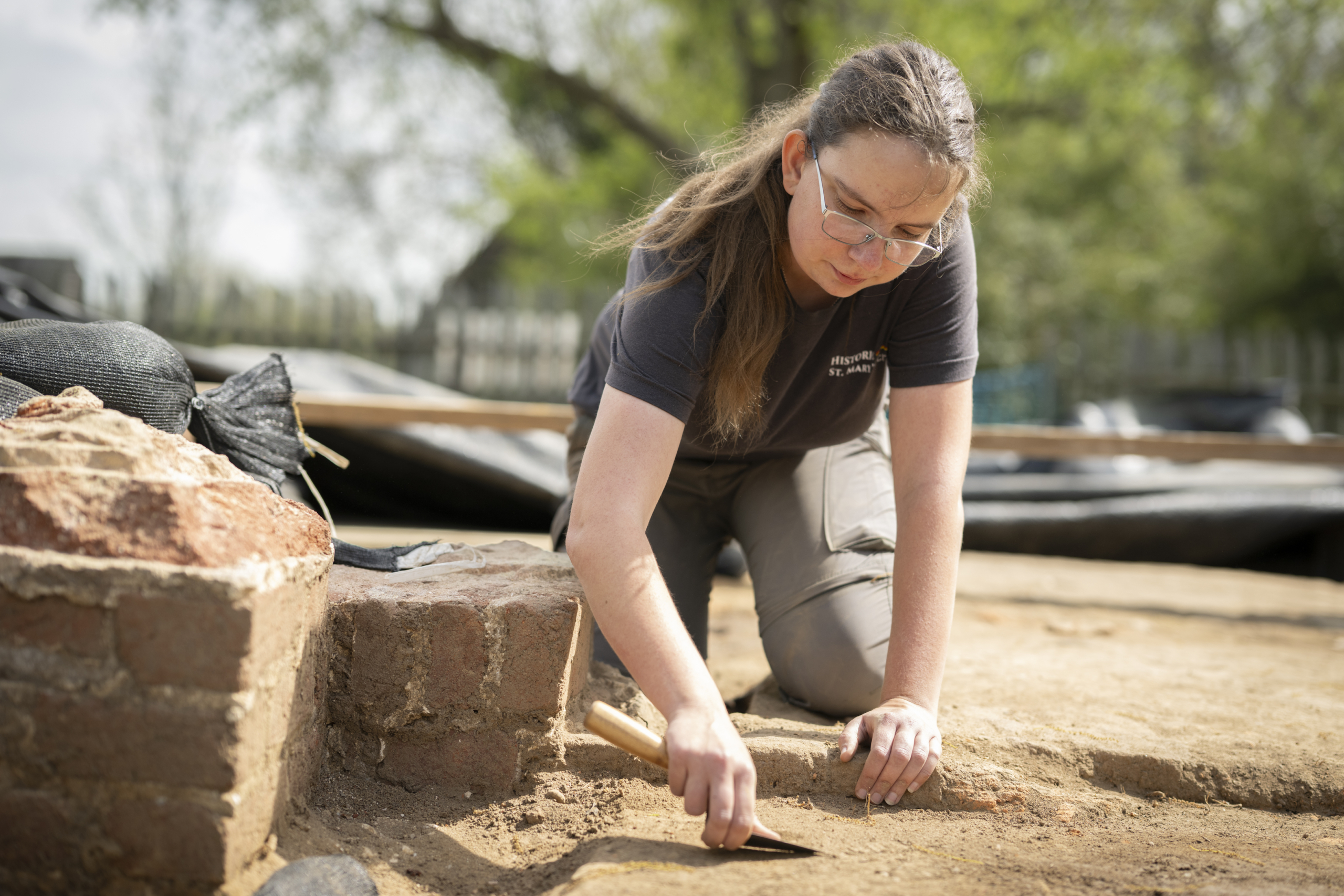 A person kneels on the ground, carefully using a small tool to excavate soil at an outdoor archaeological site.