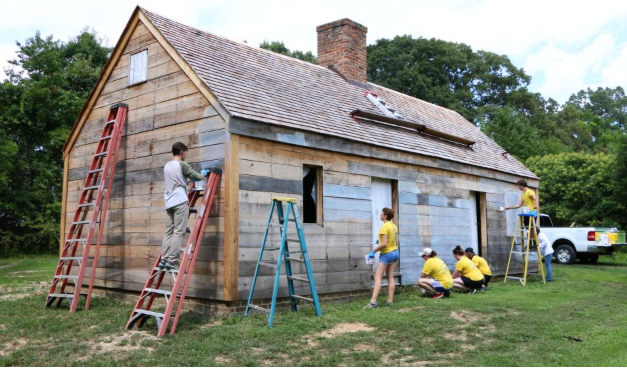 A group of people work on renovating a small wooden house, with ladders set up and some individuals painting or repairing the exterior.