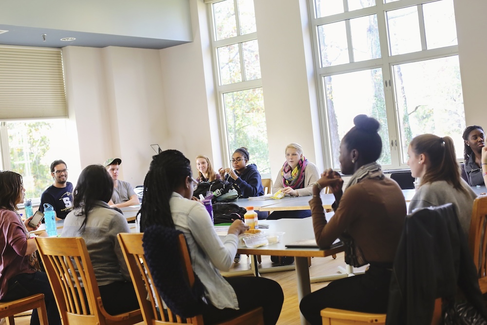 A group of people sit around a large table in a bright room with large windows, engaged in discussion.